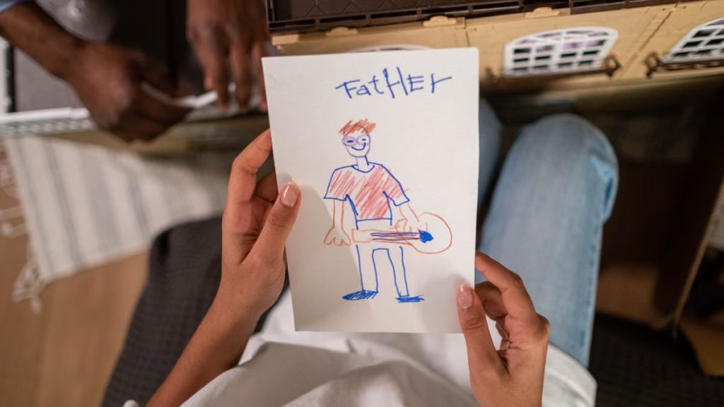 A high-angle view of a child's drawing of their father, placed in front of a toy house, showing family creativity.